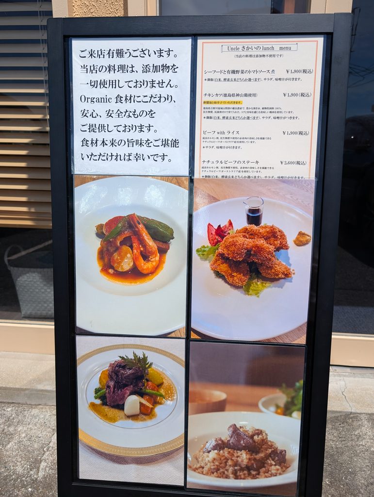 店頭の立て看板。添加物不使用の案内文とランチメニュー、料理写真4点